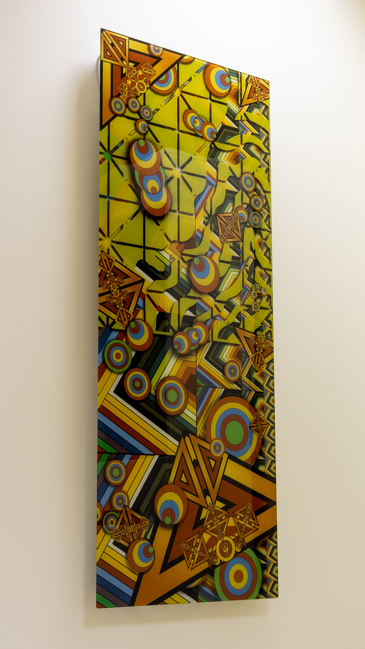 Tall wall-mounted tempered glass panel printed with colorful geometric vector art: concentric circles, chevron stripes and triangles in yellow, orange, rust, blue and green